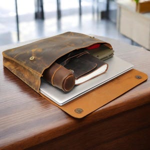 May include: A brown leather laptop bag with a flap closure, holding a silver laptop, a notebook, and a cylindrical case. The bag has a distressed finish and is open, revealing the contents. The bag is on a wooden surface.
