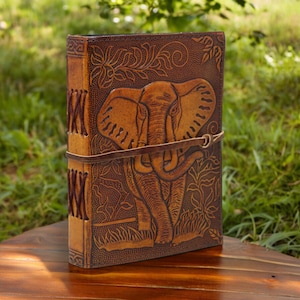 May include: A brown leather journal with an embossed elephant design and a brown leather strap closure. The journal has a decorative floral pattern around the elephant.