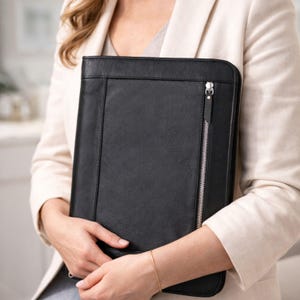 May include: A black leather portfolio with a silver zipper closure. The portfolio is rectangular and has a textured surface. The zipper runs vertically along the right side. The portfolio is held by a person wearing a cream-colored blazer.