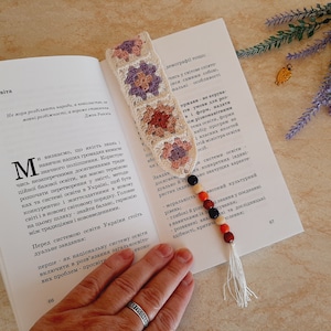 May include: A crocheted bookmark with a floral pattern in shades of pink, purple, and white. The bookmark is attached to a string of colorful beads with a tassel.