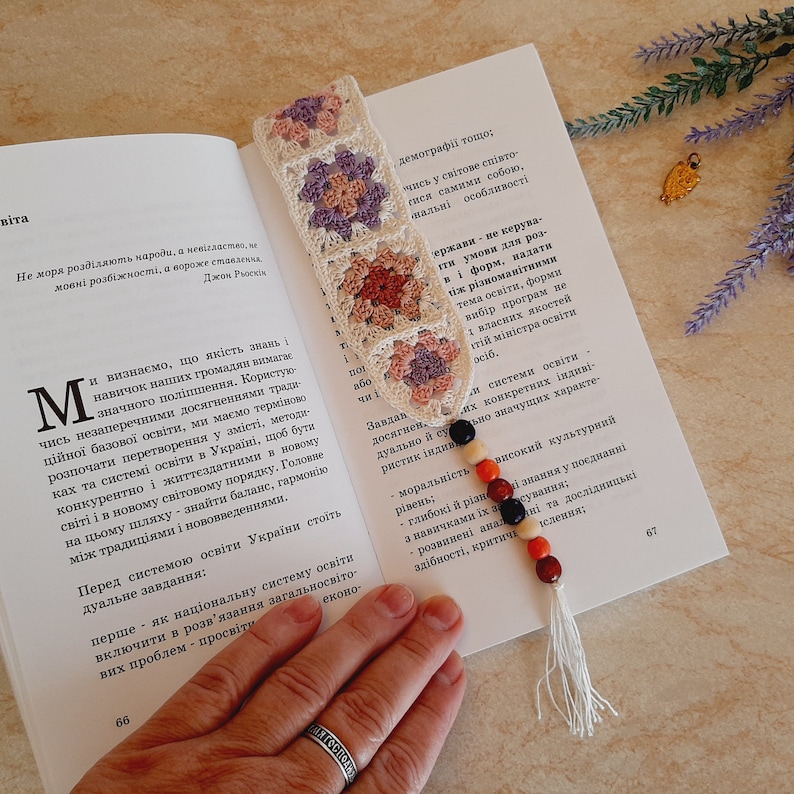 May include: A crocheted bookmark with a floral pattern in shades of pink, purple, and white. The bookmark is attached to a string of colorful beads with a tassel.