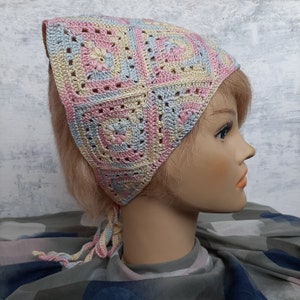 May include: A crocheted head scarf in pastel pink, blue, and yellow with a square pattern. The scarf is tied at the back of the head and has long ties that hang down the back.