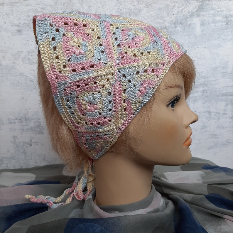 May include: A crocheted head scarf in pastel pink, blue, and yellow with a square pattern. The scarf is tied at the back of the head and has long ties that hang down the back.
