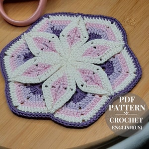 Könnte beinhalten: Ein Häkeluntersetzer mit einem Blumendesign in den Farben Lila, Rosa und Weiß. Der Untersetzer ist mit einem strukturierten Stich gearbeitet und hat einen gewellten Rand. Der Text "PDF PATTERN CROCHET ENGLISH(US)" ist auf weißem Hintergrund in der unteren rechten Ecke des Bildes gedruckt.