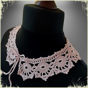 May include: A delicate pink crocheted collar with a bow tie detail. The collar is made of fine yarn and features a delicate floral pattern.
