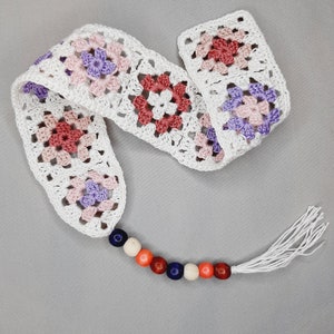 May include: White crocheted bookmark with a tassel and a string of colorful beads. The bookmark is made of granny squares with pink, purple, and red flowers.