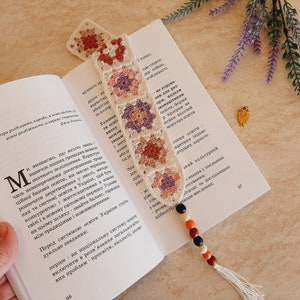 May include: A crocheted bookmark with a granny square pattern in shades of pink, purple, and cream. The bookmark is adorned with wooden beads in shades of red, orange, black, and white. The bookmark is resting on an open book with text in a foreign language.