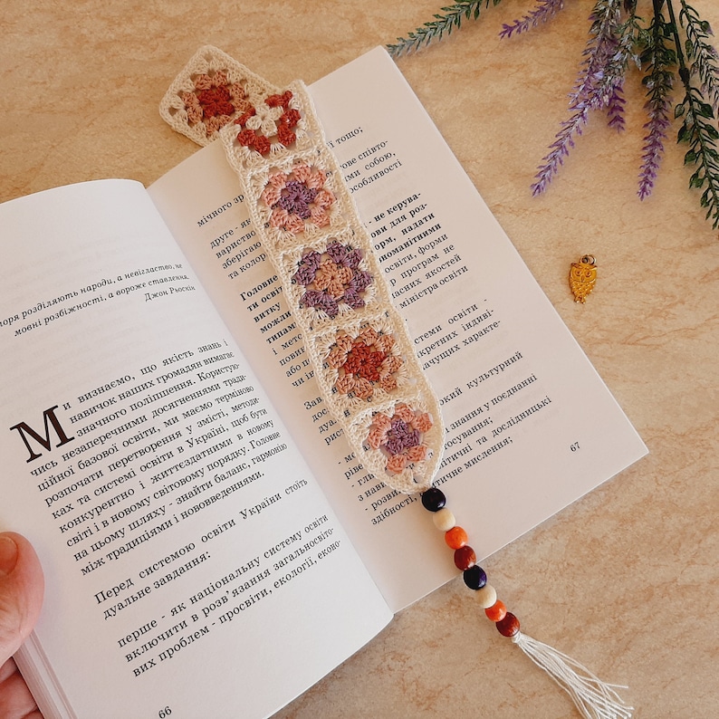May include: A crocheted bookmark with a granny square pattern in shades of pink, purple, and cream. The bookmark is adorned with wooden beads in shades of red, orange, black, and white. The bookmark is resting on an open book with text in a foreign language.