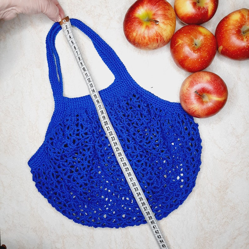 May include: A blue crocheted string bag with a white measuring tape showing the bag is 45 inches long. The bag is on a white surface with red apples around it.