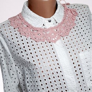 May include: A white crochet collar with a pink crocheted design. The collar is worn over a white, eyelet-patterned shirt.