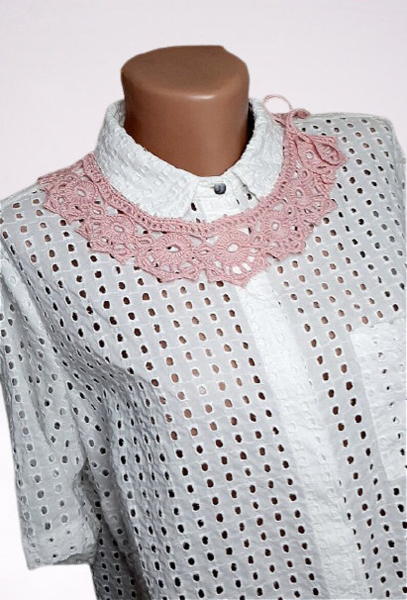 May include: A white crochet collar with a pink crocheted design. The collar is worn over a white, eyelet-patterned shirt.