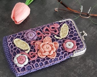 May include: A handcrafted, purple crochet glasses case with a floral design. The case features a lattice pattern and is adorned with embroidered flowers in shades of pink, peach, and lavender, with light green leaves. The case has a silver clasp.