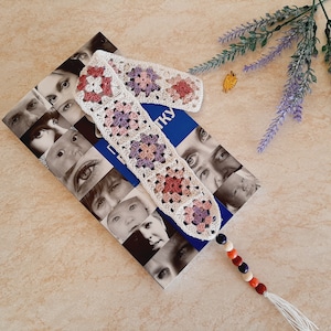 May include: A white crocheted bookmark with pink and purple squares and a tassel. The bookmark is on top of a book with a blue cover and a pattern of faces.