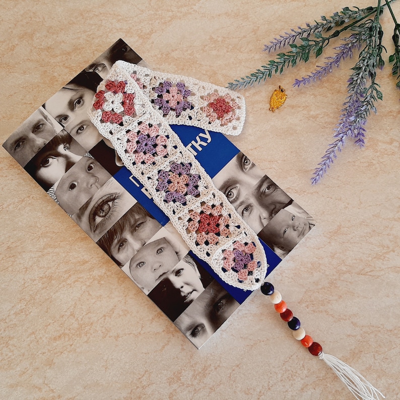 May include: A white crocheted bookmark with pink and purple squares and a tassel. The bookmark is on top of a book with a blue cover and a pattern of faces.