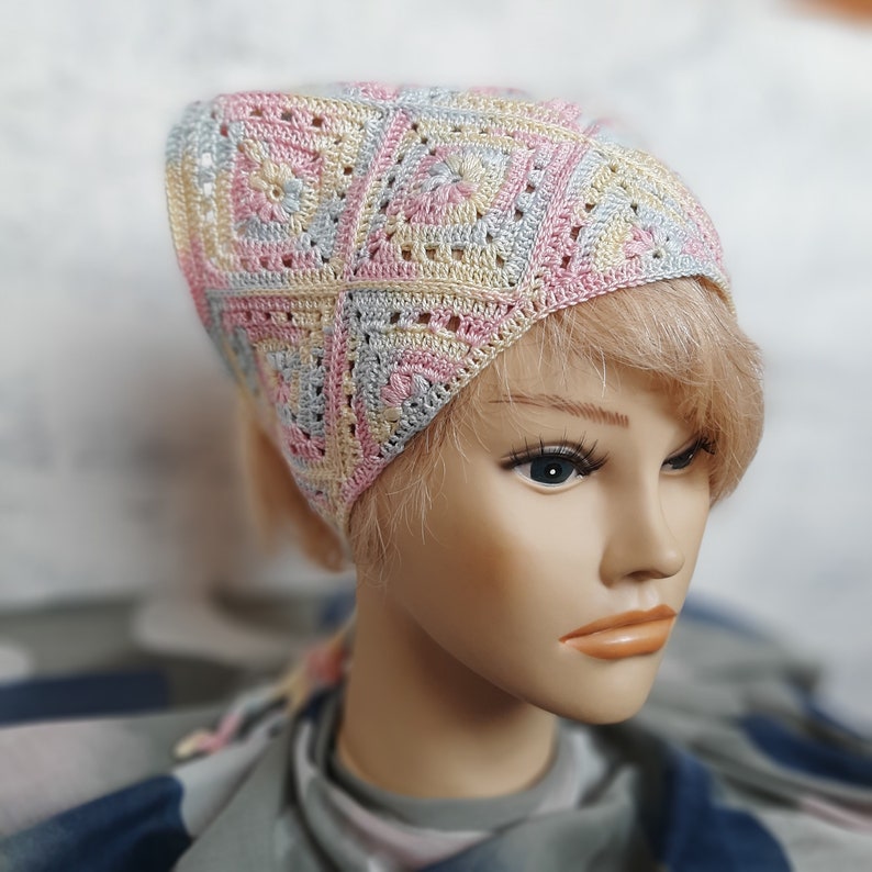 May include: A crocheted hat in pink, yellow, and gray with a square pattern. The hat is worn by a mannequin with blonde hair.