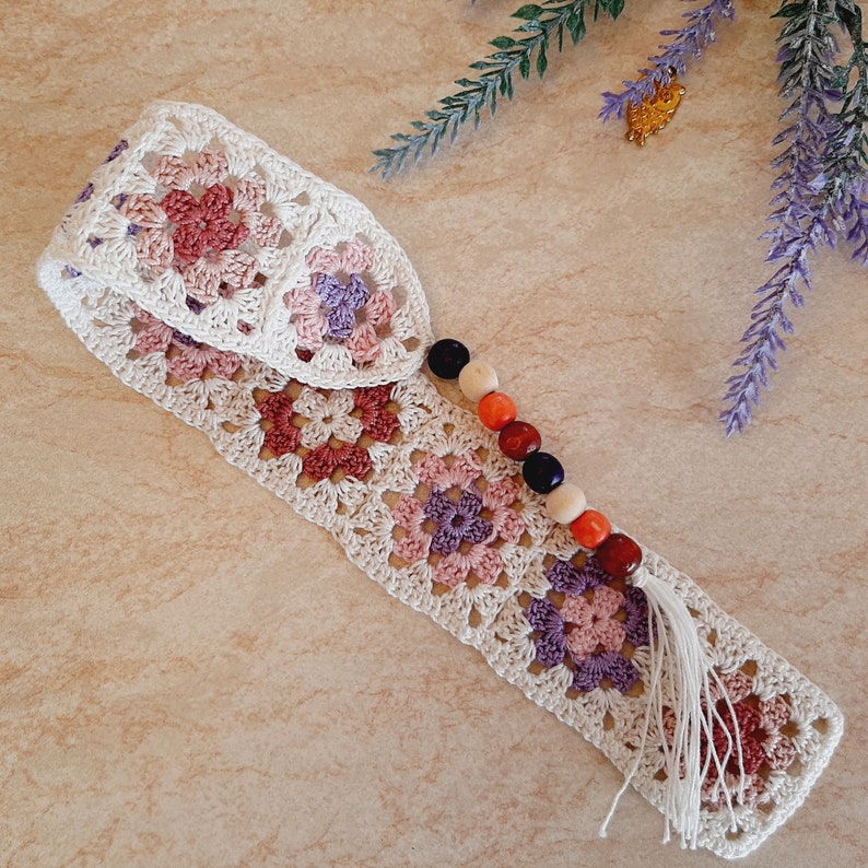 May include: A white crocheted headband with a pink, purple, and orange flower pattern. The headband has a tassel and a string of wooden beads.