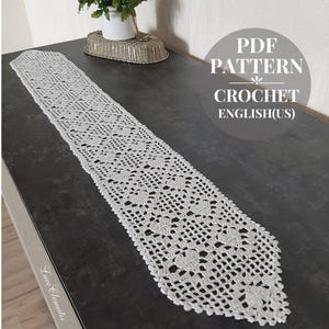May include: A white, crocheted table runner with a floral design. The runner is rectangular and is displayed on a dark surface. The image includes text that reads "PDF PATTERN CROCHET ENGLISH(US)".