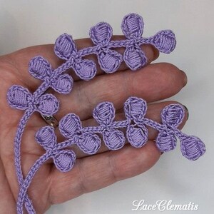May include: A close-up of a lavender crocheted lace trim with a delicate floral design. The trim features small, rounded flower shapes connected by a thin, crocheted chain. The trim is perfect for adding a touch of elegance to any project.