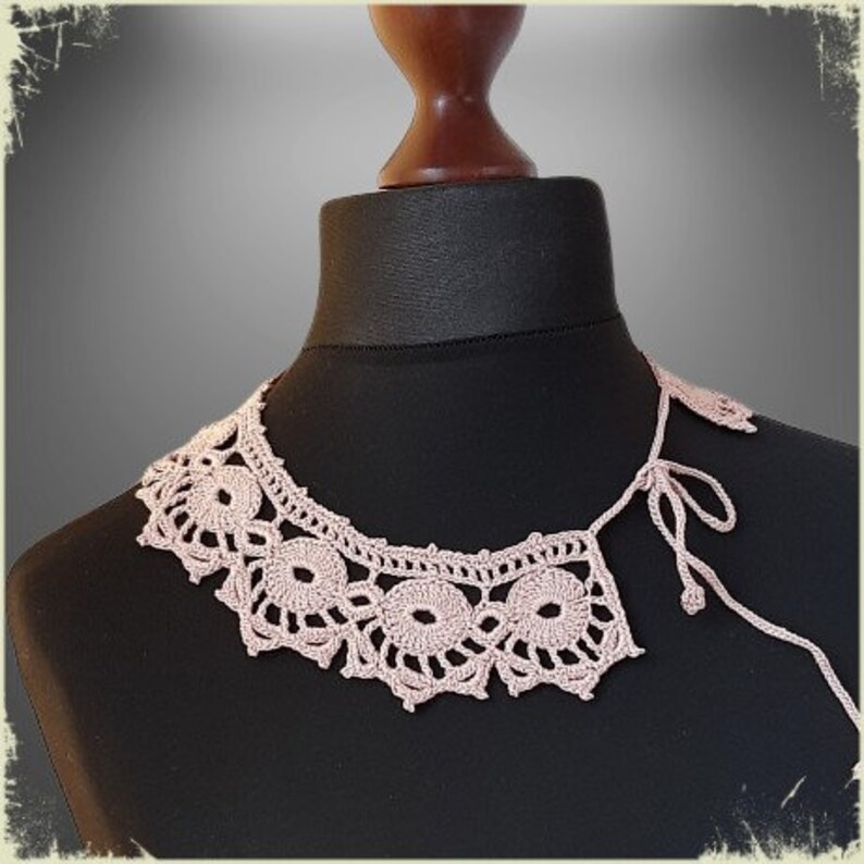 May include: A delicate pink crocheted collar with a scalloped edge. The collar is displayed on a black mannequin.