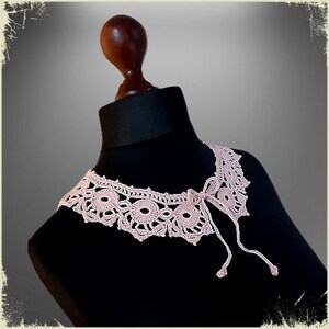 May include: A delicate pink crocheted collar with a bow detail. The collar is displayed on a black mannequin.