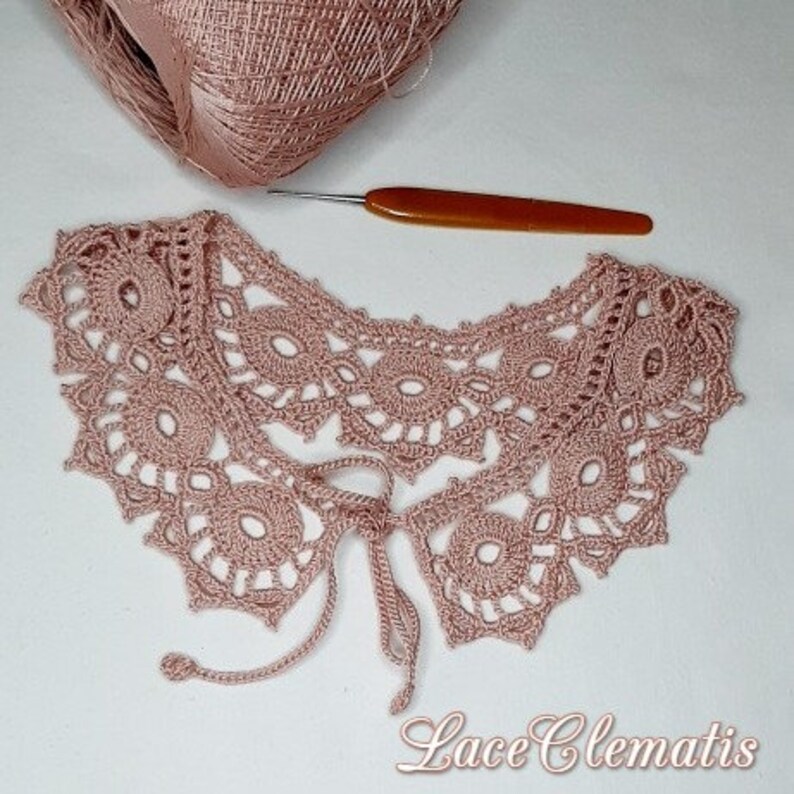 May include: A delicate pink crocheted lace collar with a tie closure. The collar is made with a fine thread and features a delicate floral pattern.