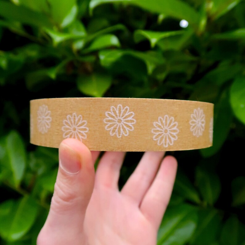 May include: A roll of brown paper tape with a white daisy pattern. The tape is being held in a hand.