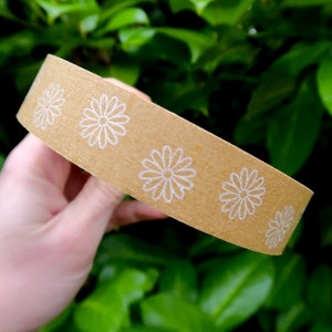 May include: A roll of brown packing tape with a white daisy pattern. The tape is being held in a hand.