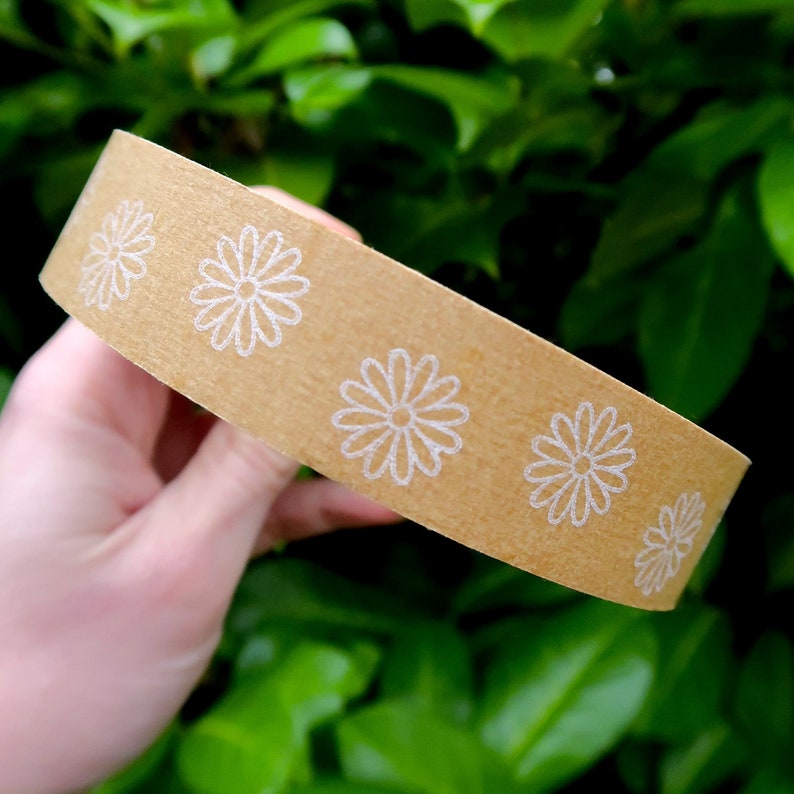 May include: A roll of brown packing tape with a white daisy pattern. The tape is being held in a hand.