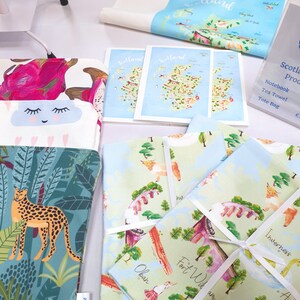 May include: A collection of tea towels featuring illustrated maps of Scotland, including Oban, Fort William, and Inverness. The tea towels are folded and tied with white ribbon. The tea towels are on a white surface.