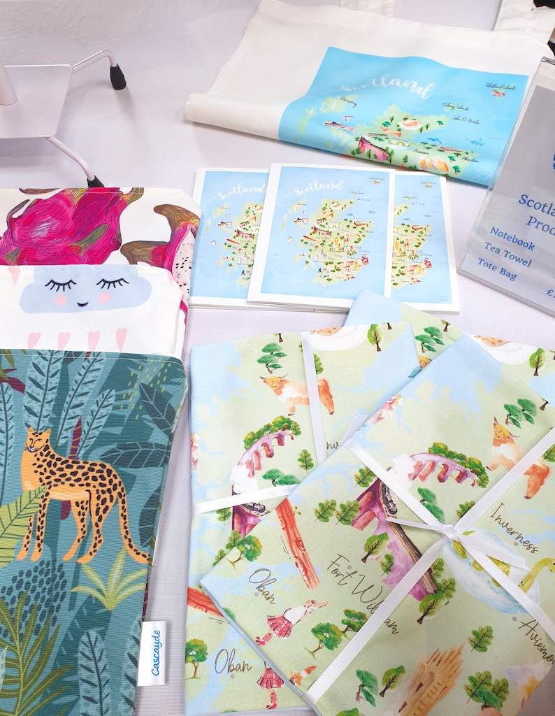 May include: A collection of tea towels featuring illustrated maps of Scotland, including Oban, Fort William, and Inverness. The tea towels are folded and tied with white ribbon. The tea towels are on a white surface.