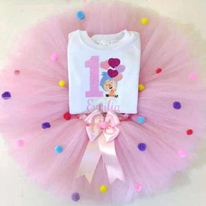Può includere: Un tutù rosa con pompon colorati e una maglietta bianca. La maglietta ha un grande numero 1 in glitter rosa, un personaggio dei cartoni animati e il nome "Emilia". Un fiocco rosa è in vita.