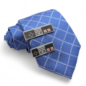 May include: A blue tie with a diamond pattern, paired with cufflinks designed as retro video game controllers. The cufflinks are gray and black with red and white accents, adding a playful touch to the formal accessory.