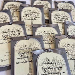 May include: Gray seed packets with a tree design and the text "Those we love don't go away, they walk beside us every day." Each packet is tied with a gray ribbon. The packets are for memorial gifts.