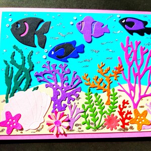 May include: A pink card with a blue background featuring a variety of colorful cut-out shapes, including fish, coral, and seaweed. The card has a pink border and a white seashell in the lower left corner.