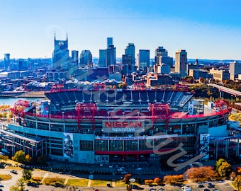 Może przedstawiać: Widok lotniczy na Nissan Stadium w Nashville w stanie Tennessee, z panoramą miasta w tle. Stadion jest dużą, otwartą konstrukcją z czerwonymi i niebieskimi siedzeniami. Stadion jest otoczony drzewami i parkingami.