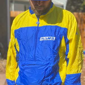 Puede incluir: Chaqueta cortavientos amarilla y azul de los Rams con cierre de media cremallera y la palabra "RAMS" impresa en la parte delantera. La chaqueta está hecha de un material ligero y tiene un bolsillo azul.