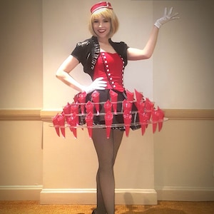 May include: A woman wearing a red and black costume with a skirt made of red cones. She is wearing a black jacket, a red corset, and a black hat with a red band. She is standing on a wooden floor and has her right hand raised.