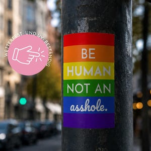 Könnte beinhalten: Ein rechteckiger Aufkleber mit Regenbogenhintergrund und dem weißen Text BE HUMAN NOT AN asshole. Links befindet sich ein kreisförmiges rosa Abzeichen mit einem weißen Hand-Symbol, das nach rechts zeigt, sowie dem Text WETTERFEST UND UV-BESTÄNDIG.