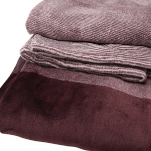 May include: A folded textile blanket featuring a deep burgundy velvet-like border and a textured, heathered maroon and white woven fabric section. The material displays a distinct contrast between the smooth, solid dark red edge and the ribbed pattern.