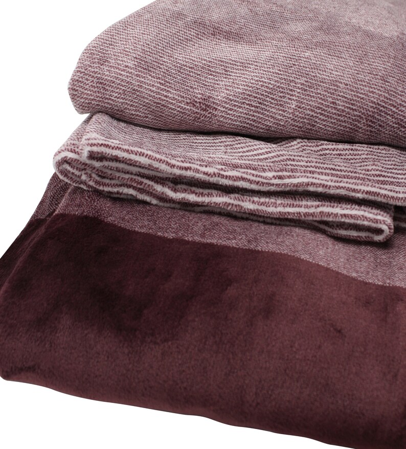 May include: A folded textile blanket featuring a deep burgundy velvet-like border and a textured, heathered maroon and white woven fabric section. The material displays a distinct contrast between the smooth, solid dark red edge and the ribbed pattern.
