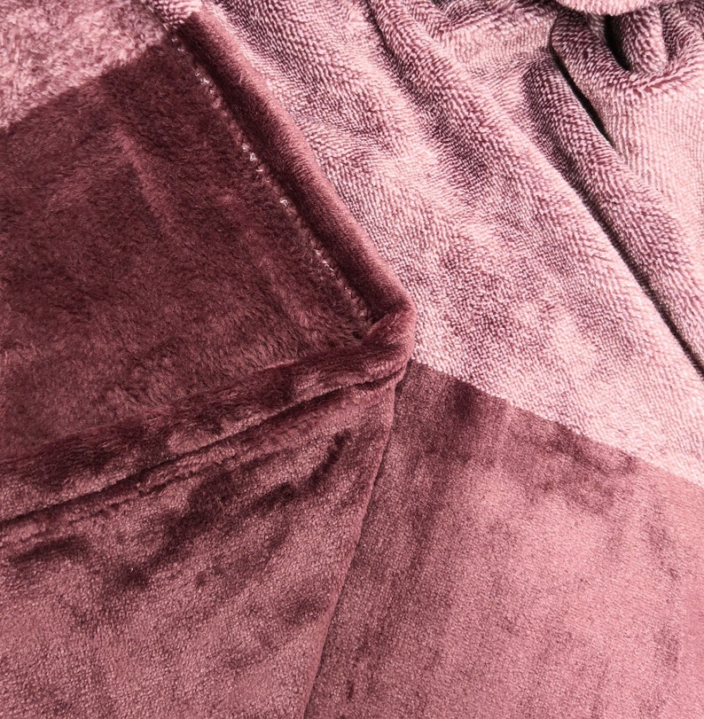 May include: A plush, mauve-colored blanket featuring a soft, velvety texture on one side and a looped, terry-cloth-like fabric on the other. The material is folded to reveal the contrast between the smooth, dense pile and the textured weave.