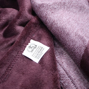 May include: A deep plum-colored plush blanket with a soft, fuzzy texture. A white fabric care label is stitched onto the corner, featuring the text Henry's, 100% Polyester, Wash with similar colors, and Do not ask your wife or mom.
