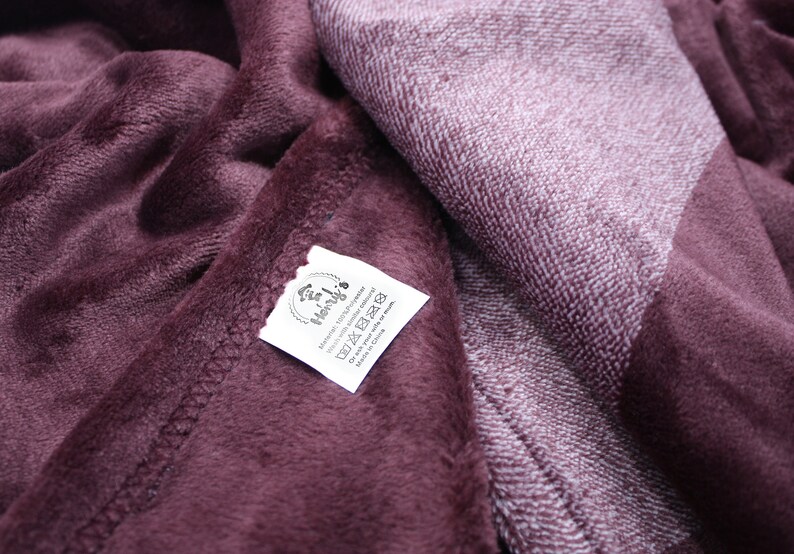 May include: A deep plum-colored plush blanket with a soft, fuzzy texture. A white fabric care label is stitched onto the corner, featuring the text Henry's, 100% Polyester, Wash with similar colors, and Do not ask your wife or mom.
