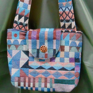 May include: A colorful woven shoulder bag with a geometric pattern in shades of blue, pink, and brown. The bag has a flap closure with a wooden button.
