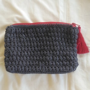 May include: A gray crocheted pouch with a red zipper and tassel. The pouch has a textured, bumpy surface.