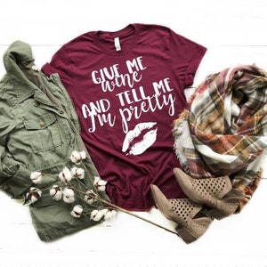 May include: Burgundy t-shirt with white text that reads "Give me wine and tell me I'm pretty". The shirt features a white lip print. The shirt is laid out on a white surface with a green jacket, a plaid scarf, and brown boots.