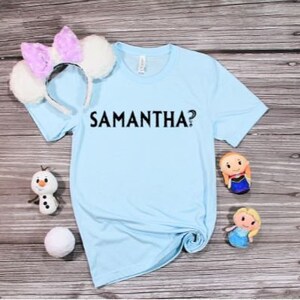 Puede incluir: Una camiseta azul claro con el texto "SAMANTHA?" impreso en negro. La camiseta está sobre una superficie de madera con una diadema blanca, una figura de muñeco de nieve y dos muñecas pequeñas.