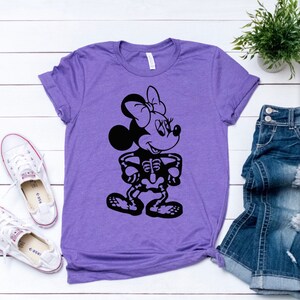 May include: Purple t-shirt with a black graphic of a skeleton Mickey Mouse character. The character is wearing a bow and has a smile.