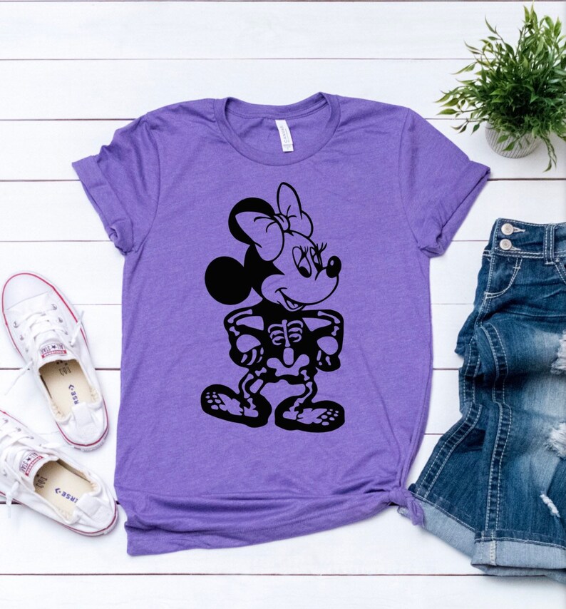 May include: Purple t-shirt with a black graphic of a skeleton Mickey Mouse character. The character is wearing a bow and has a smile.