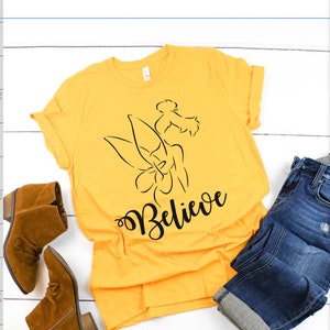 Puede incluir: Una camiseta amarilla con un contorno negro de Campanilla y la palabra "Believe" en letra negra.